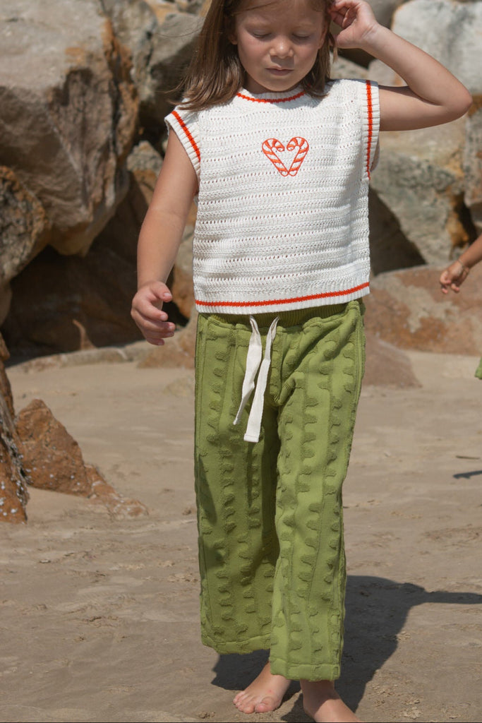 Child wearing a white knit vest with red candy canes and green towelling pants standing on a stone surface.