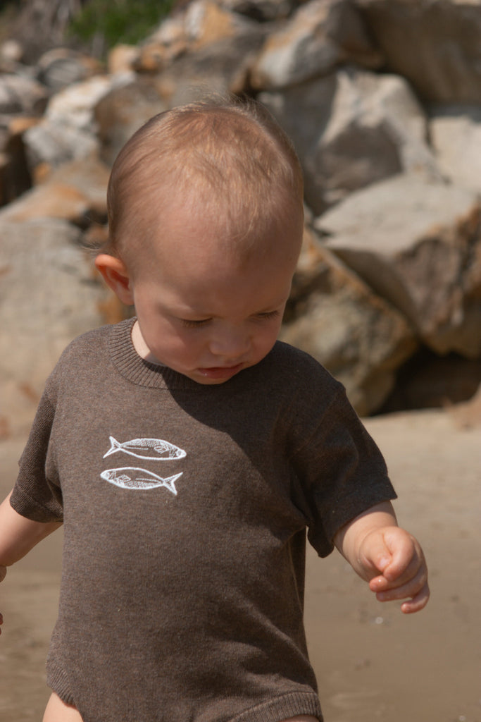 Child wearing a brown romper with fish designs, standing on a rocky beach.