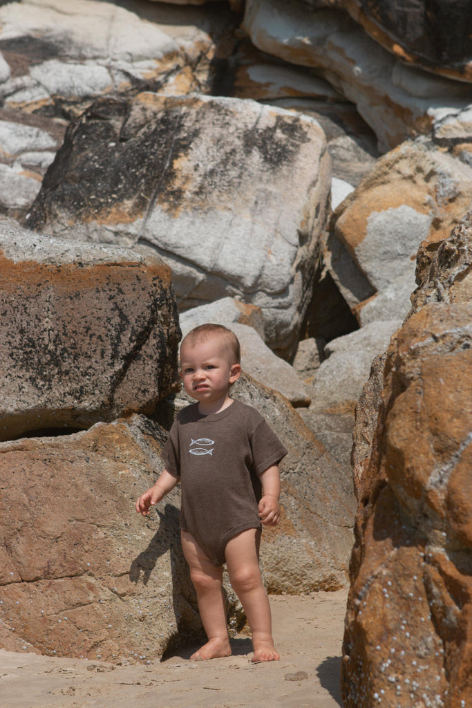 Child in a brown onesie standing among large rocks on a beach
