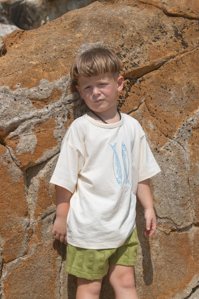 Child wearing a white t-shirt with blue fish and green towelling shorts standing against a rocky wall.