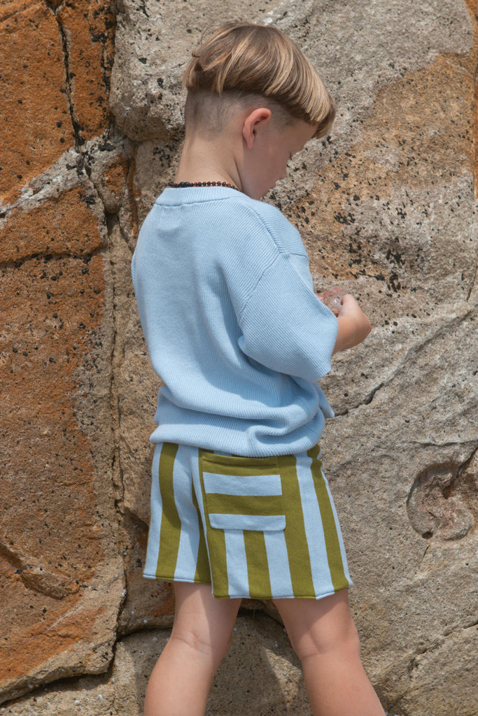 Child wearing a light blue shirt and striped shorts against a stone wall.