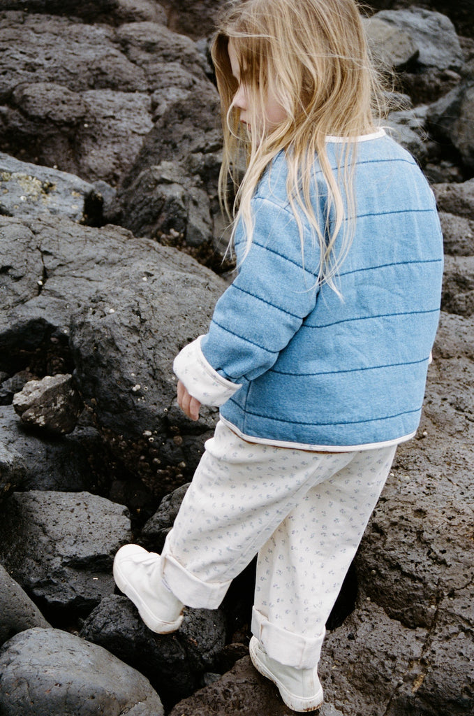 Child wearing a blue quilted jacket and white cotton floral pants standing on rocky terrain