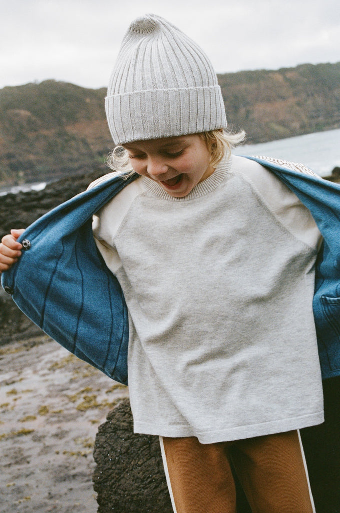 Child wearing a gray knit hat and sweater with a blue coat, standing outdoors by a body of water.