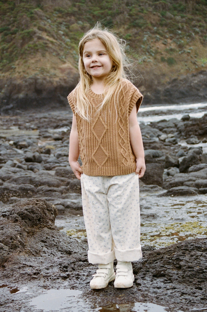 child wearing a brown knit vest and white cotton floral pants  on a rocky beach