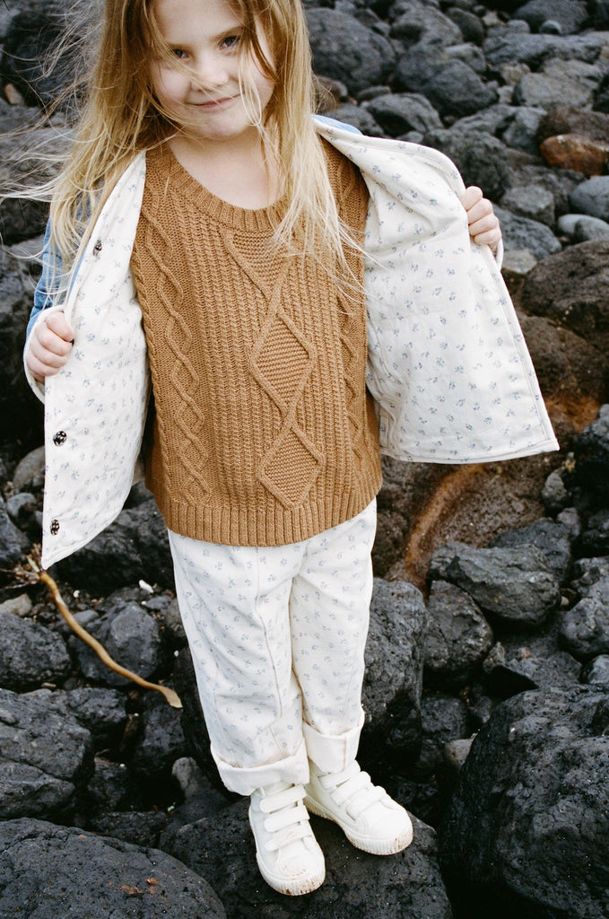 child wearing a brown knit vest and white cotton floral pants  on a rocky beach