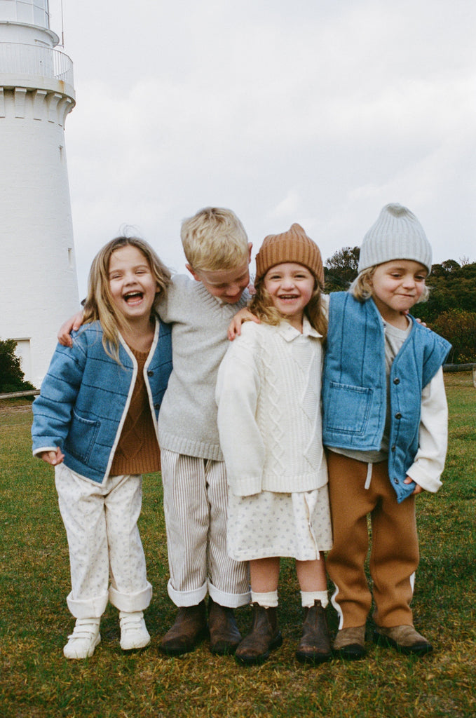 Four children standing together in a grassy area with a lighthouse in the background.
