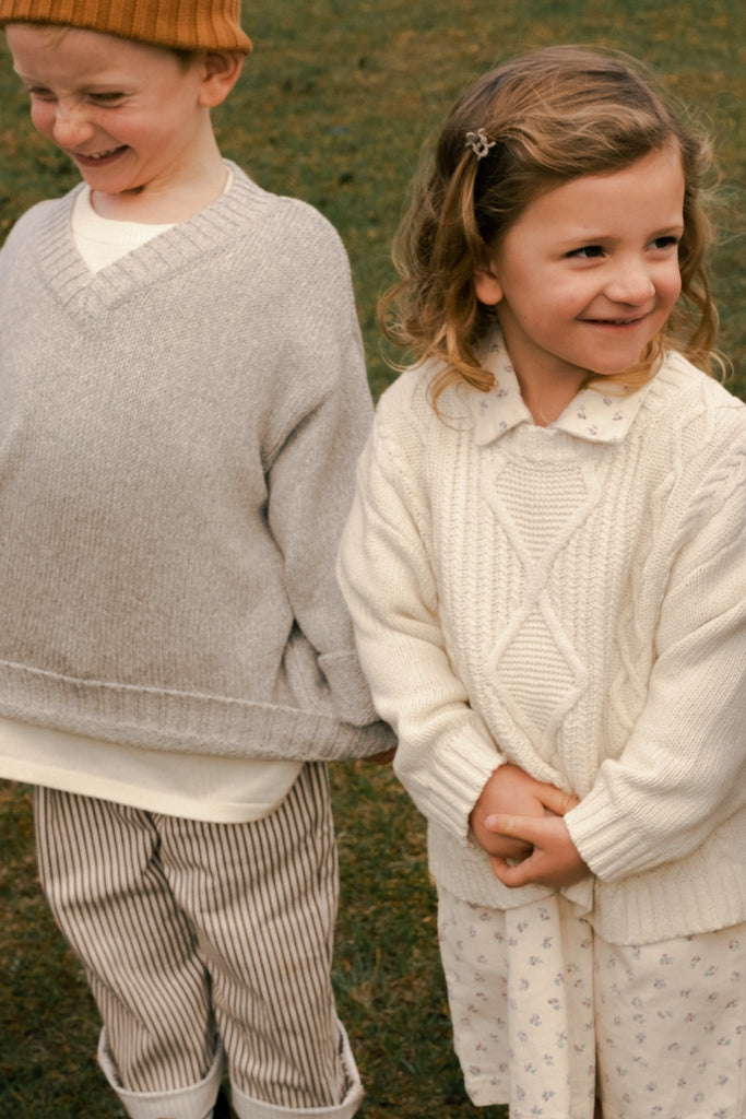 Two children wearing knitted sweaters standing outdoors on a grassy field.