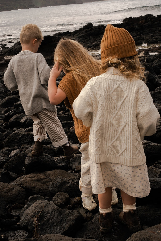 Three children on a rocky beach wearing knitted clothing.