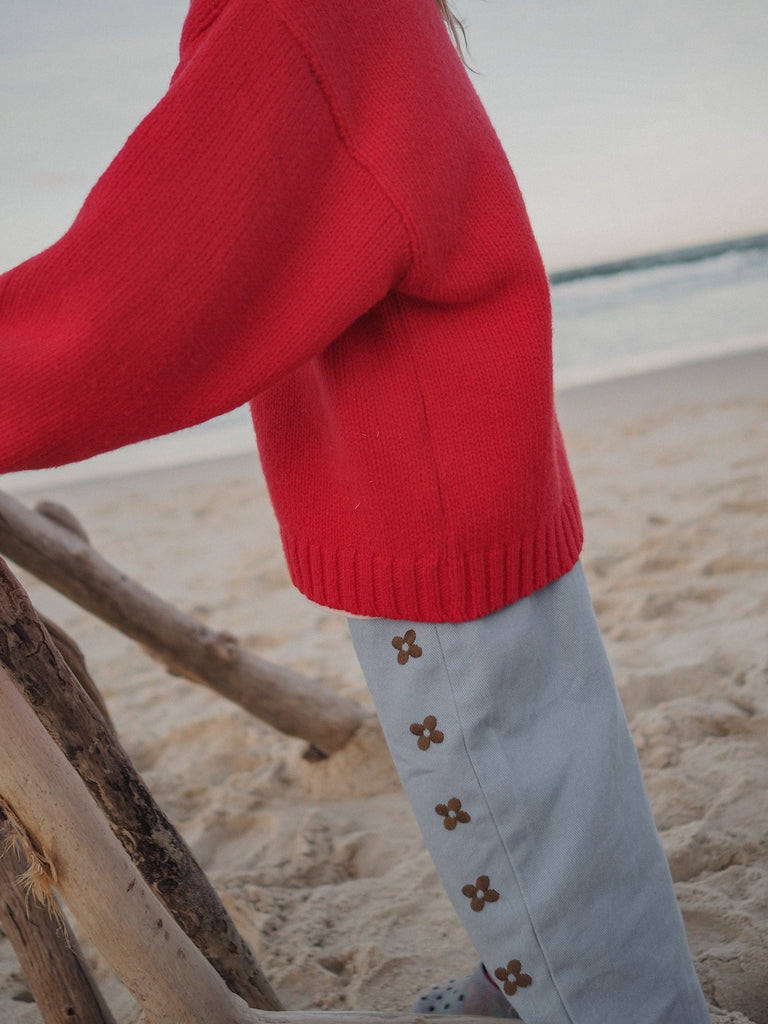 Red sweater and light denim blue pants with floral embroidery on a beach.