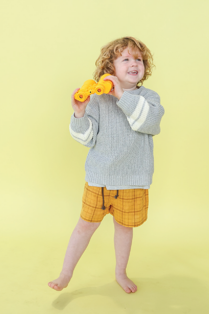 Child wearing grey knit jumper and yellow check shorts n a yellow background