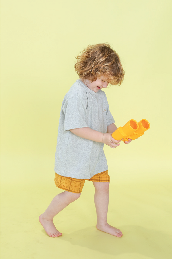 Child wearing a grey shirt and yellow shorts, holding toy binoculars against a yellow background