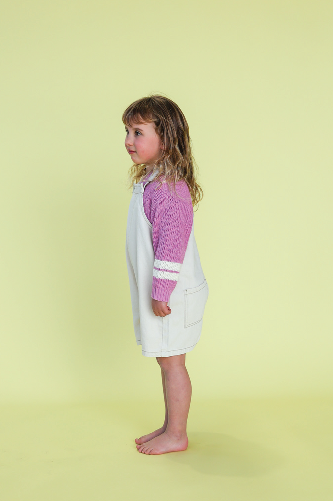 Young girl wearing a white overalls with pink knit jumper against a yellow background