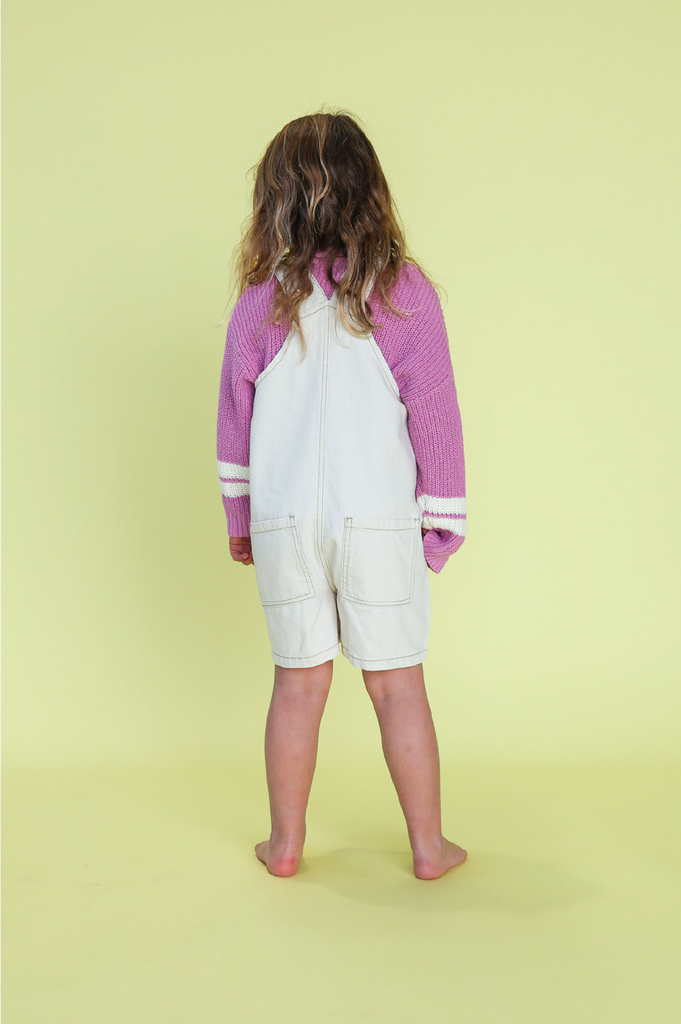 Young girl wearing a white overalls with pink knit jumper against a yellow background