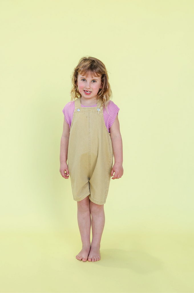 Young girl wearing mustard overalls and a pink knit vest against a yellow background