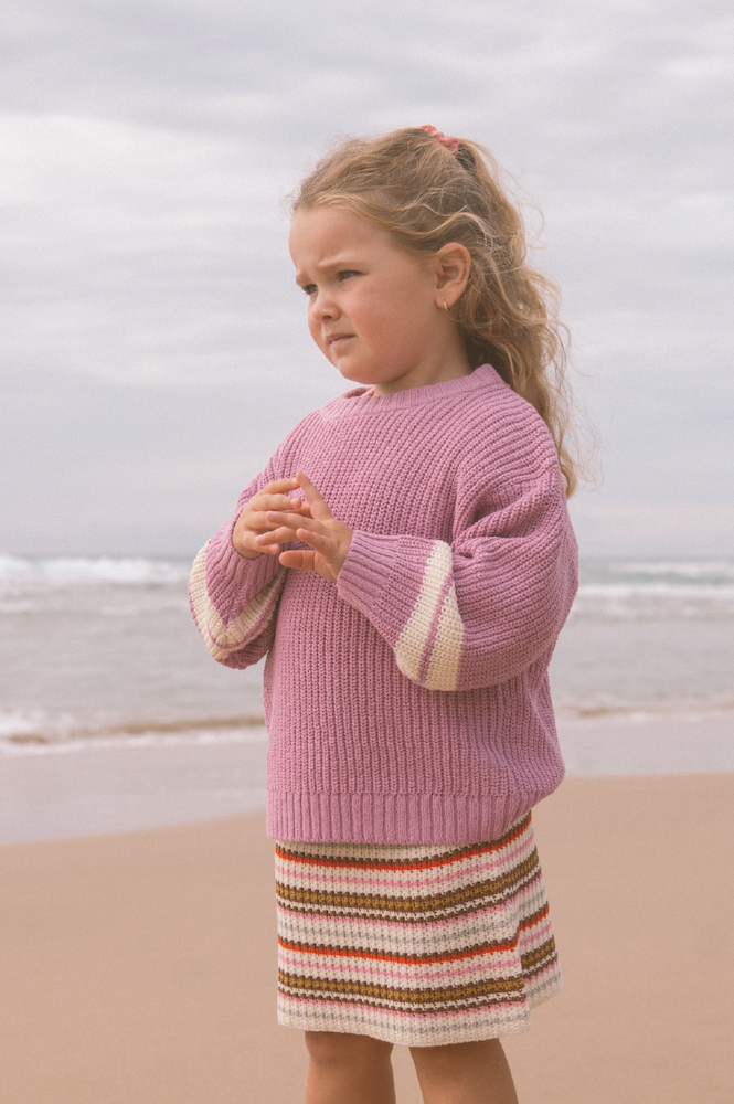 Young girl wearing a pink knitted jumper and striped skirt standing on a beach.
