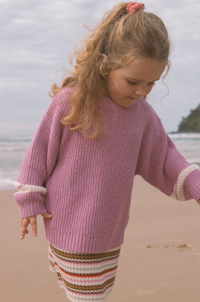 Young girl wearing a pink knitted jumper and striped skirt standing on a beach.