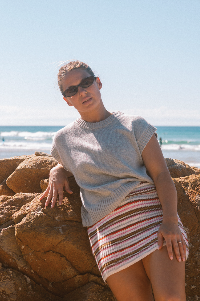 Person wearing a grey knit vest and striped skirt standing on a beach.