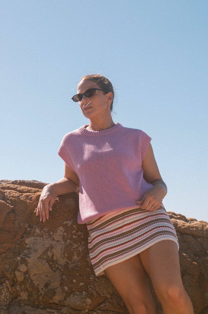 Person wearing a pink knit vest and striped skirt standing on a beach.