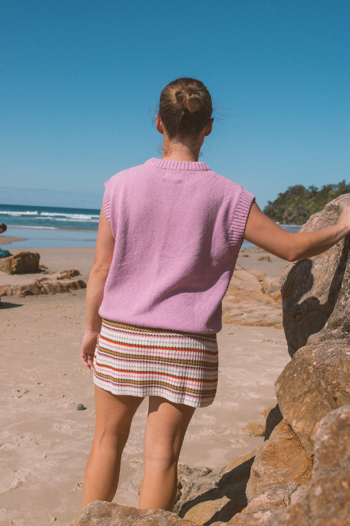 Person wearing a pink knit vest and striped skirt standing on a beach.