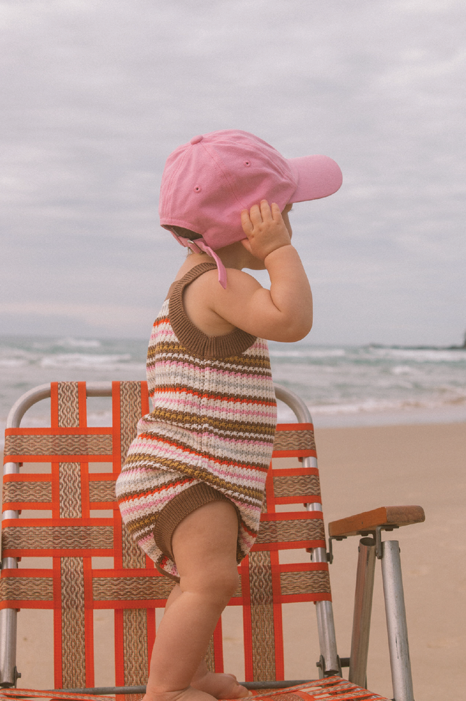Child wearing a pink cap and striped romper standing on a beach chair at the beach.