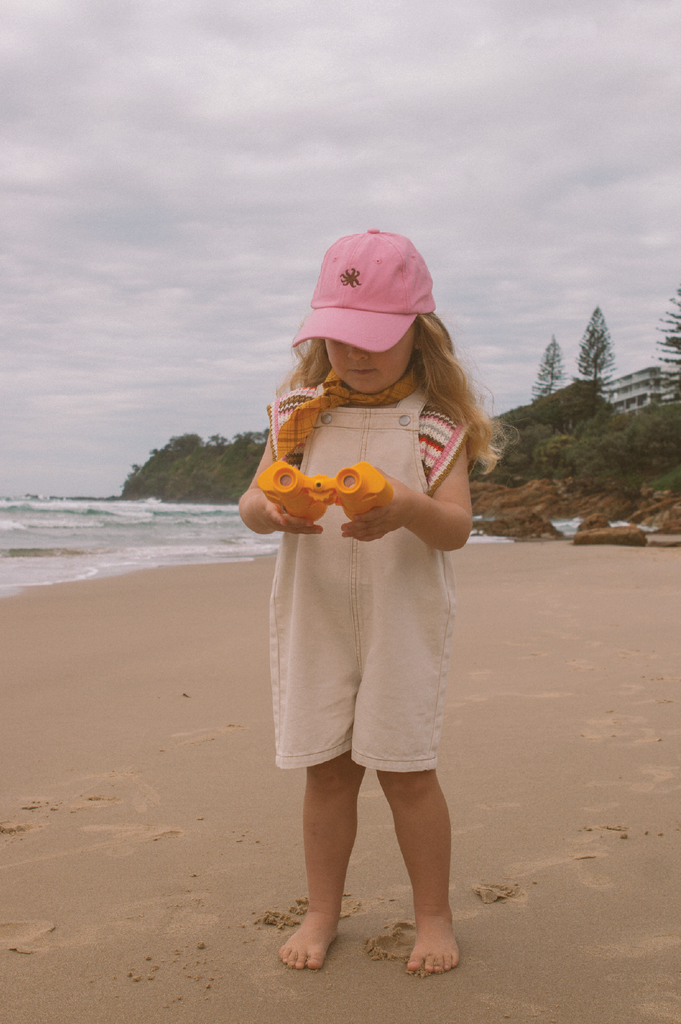 Child on a beach wearing a pink cap and white denim overalls