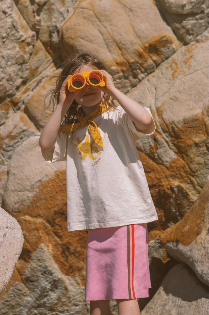 Child playing with toy binoculars against a rocky background wearing a pink skirt and white tee