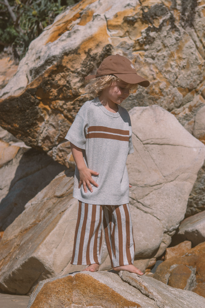 Young boy wearing a grey and brown t-shirt and brown and grey stripe pants standing on a rock