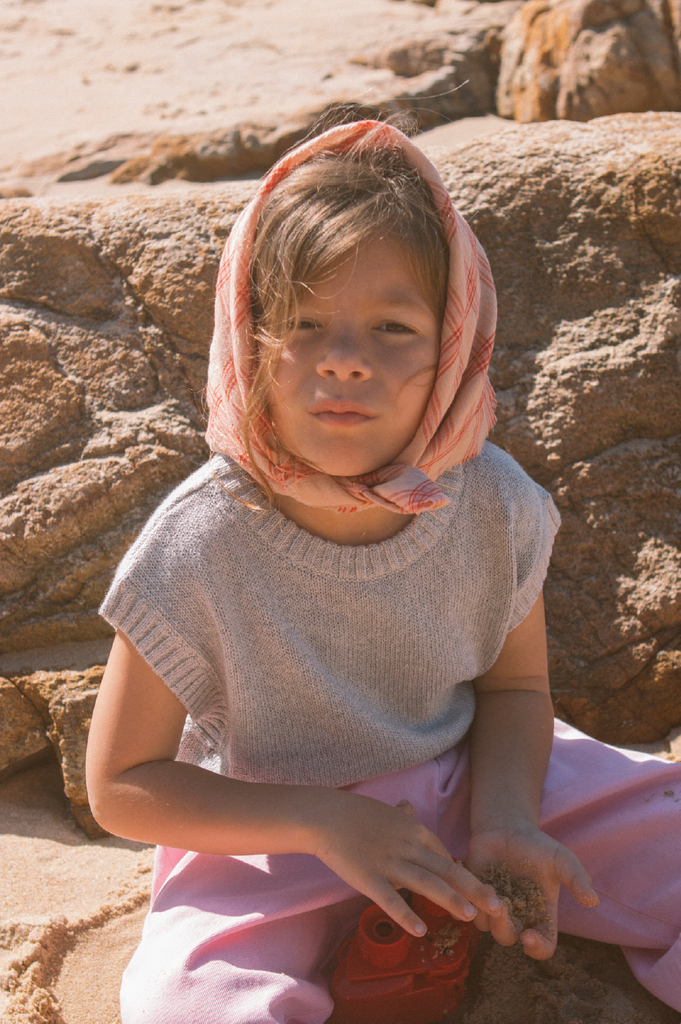 Young girl sitting on rocks wearing a pink headscarf and pink pants and grey knit vest