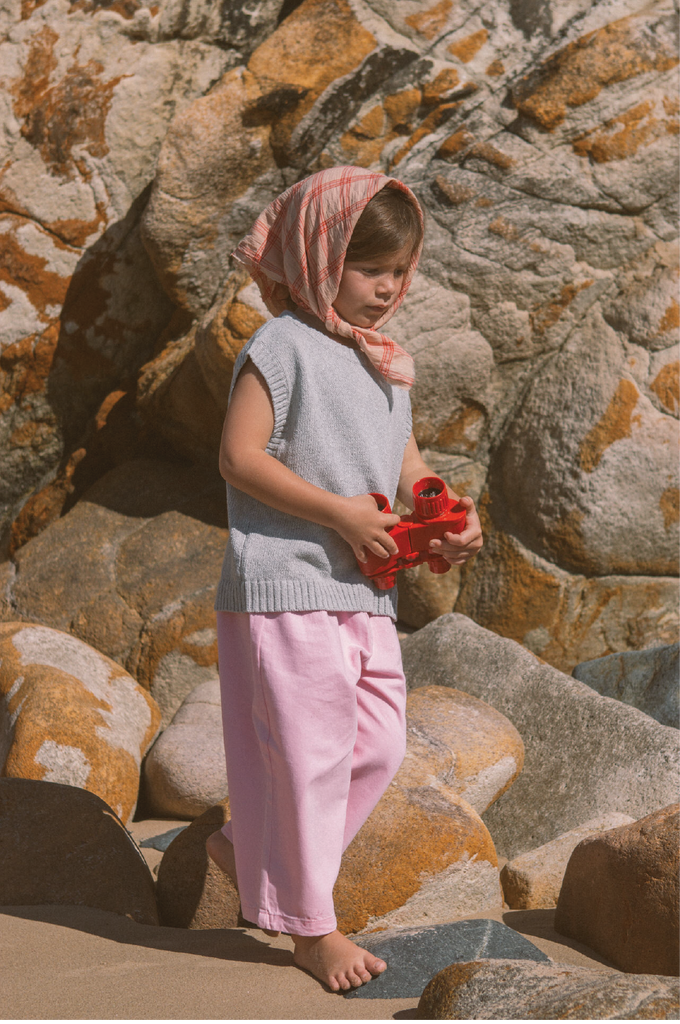 Child wearing a pink headscarf, grey vest shirt and pink denim jeans standing on rocks with a stone wall background