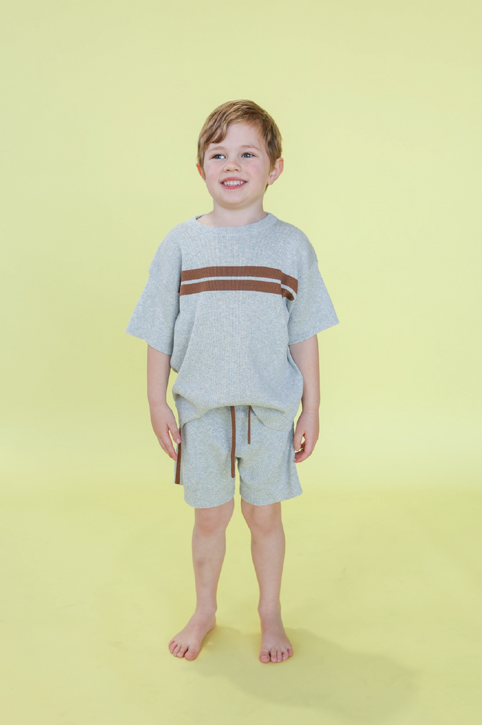 Child wearing a grey t-shirt with brown stripes and grey stripe shorts on a yellow background