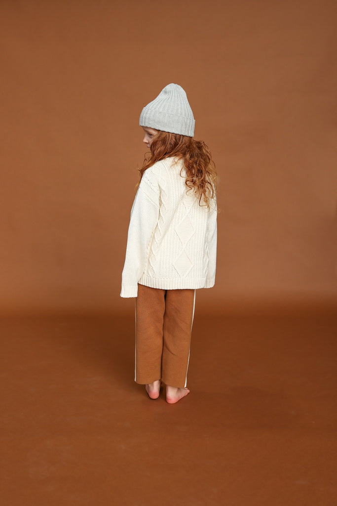 White milk sweater with cable knit pattern made from organic cotton for children with grey knitted beanie and brown knitted pants back veiw