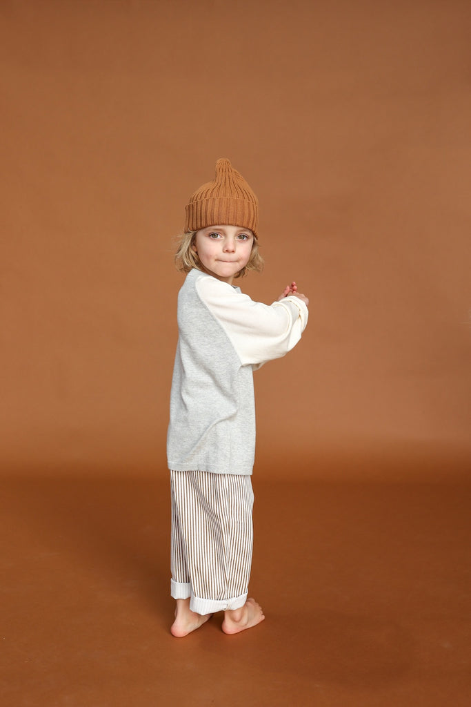 Child wearing a gray sweater and striped pants with a brown beanie against a brown background