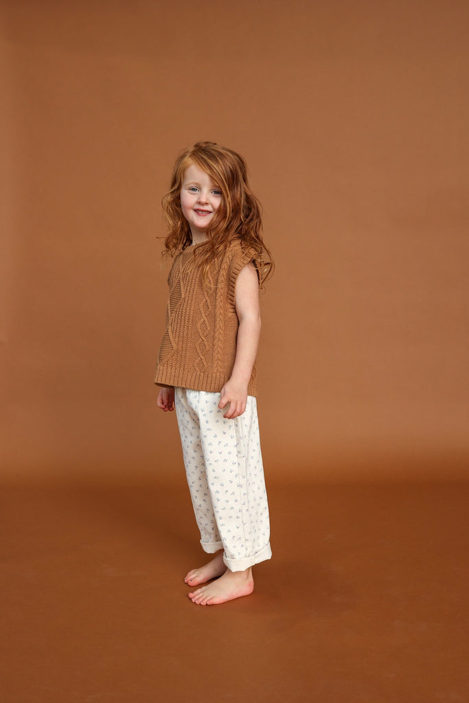 child wearing a brown knit vest and white cotton floral pants against a brown background