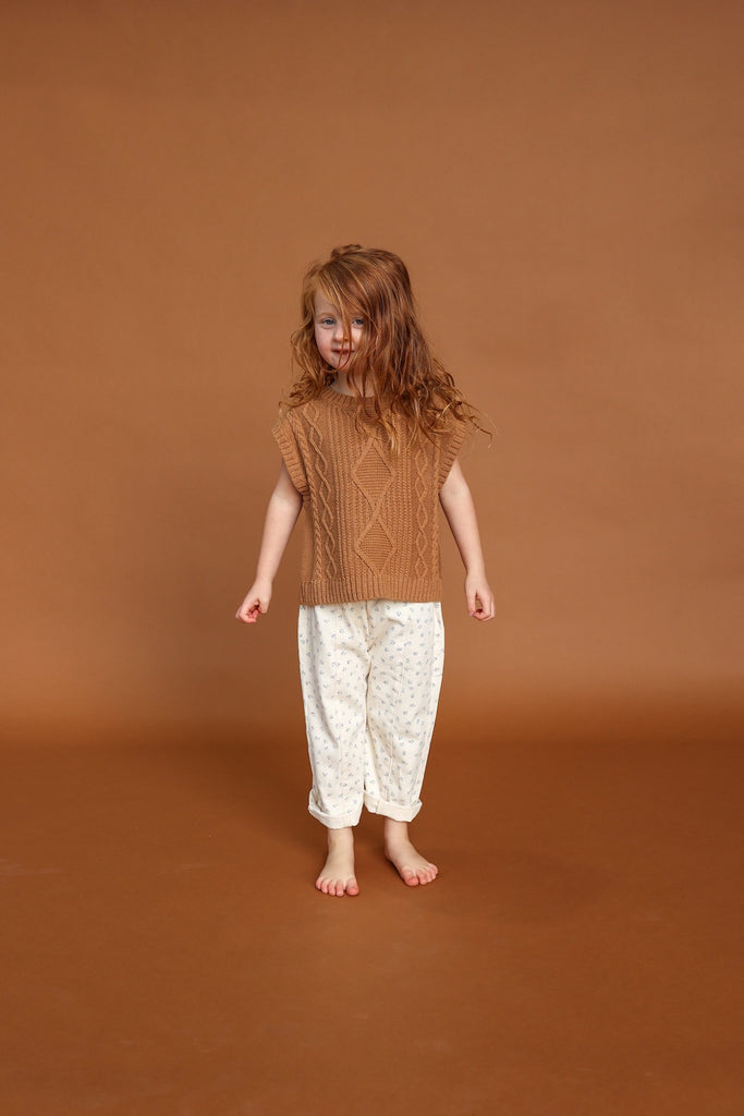 Child wearing a brown knitted top and white floral pants on a brown background