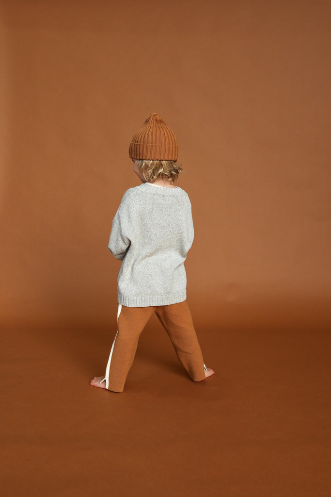 Child wearing a light gray sweater and brown pants standing against a brown background