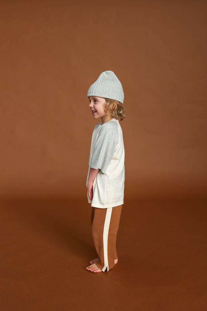 Child wearing a light gray knit hat and a neutral coloured outfit on a brown background, side view